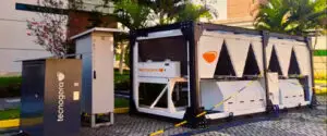 Gerador branco da Tecnogera ao ar livre, próximo a um quadro elétrico, entre plantas e palmeiras; acesso isolado por fita amarela.