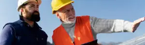 No canteiro de obras, dois homens usam capacetes. Um escuta atento enquanto o outro aponta algo à frente. Tecnologia ao fundo.