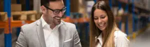 Na imagem do portal Tecnogera, um homem de terno cinza claro e uma mulher sorridente observam juntos algo no corredor do almoxarifado.
